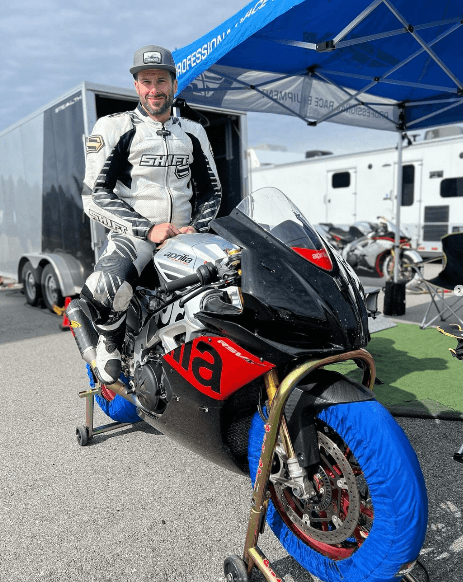 Man in motorcycle gear posing on an Aprilia bike under a blue canopy at a racetrack.